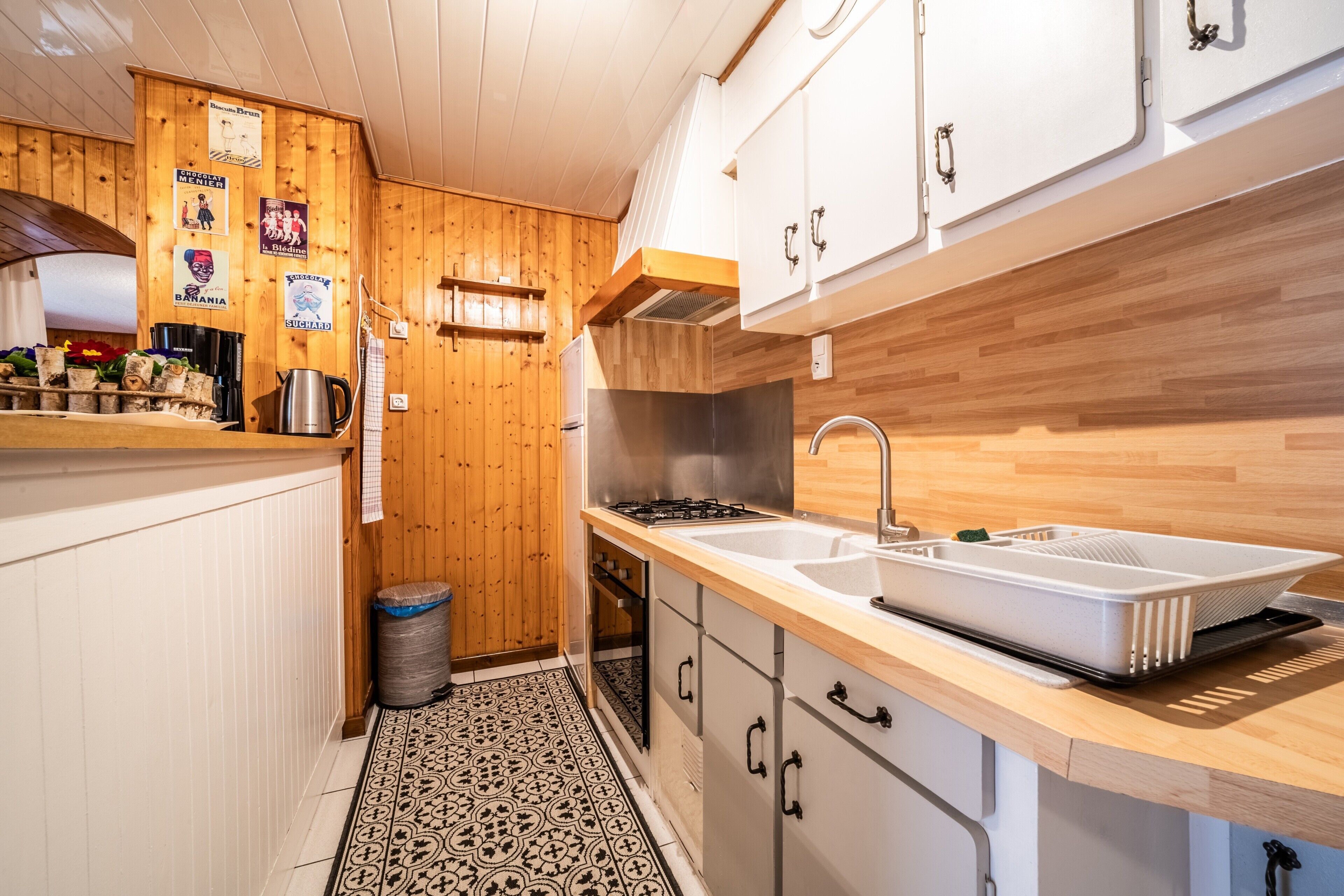 Een keuken met houten wanden, witte kasten, een roestvrijstalen spoelbak en een betegelde vloer.