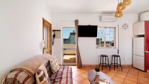 Un espace de vie compact comprenant un canapé, une petite table à manger, deux tabourets, un téléviseur et un coin cuisine avec un réfrigérateur et des placards.