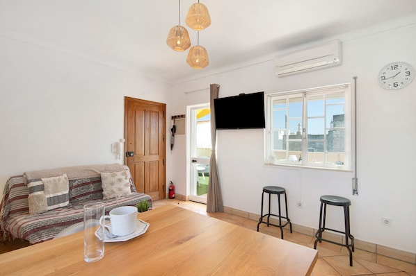 A room with a wooden table, a sofa, two bar stools, a TV, and a window with a view.
