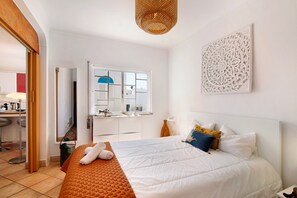 A bedroom with a bed, a window, a mirror, a wall art piece, and a door leading to another room.
