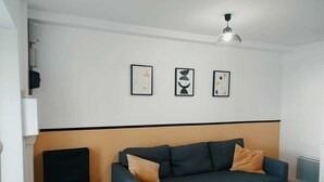 Een woonkamer met een donkergroene bank, twee oranje kussens en drie abstracte kunstwerken aan de muur.