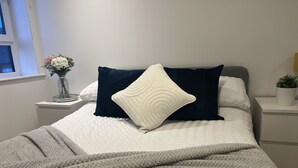 A neatly made bed with a white and navy blue pillow arrangement, a grey blanket, and a bedside table with a vase of flowers. -  Hull Spacious Apartment 7 (Hull)