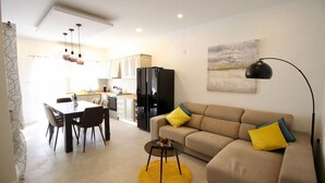 A modern living room with a sofa, dining table, and chairs. There is a kitchen area in the background with cabinets and a refrigerator.