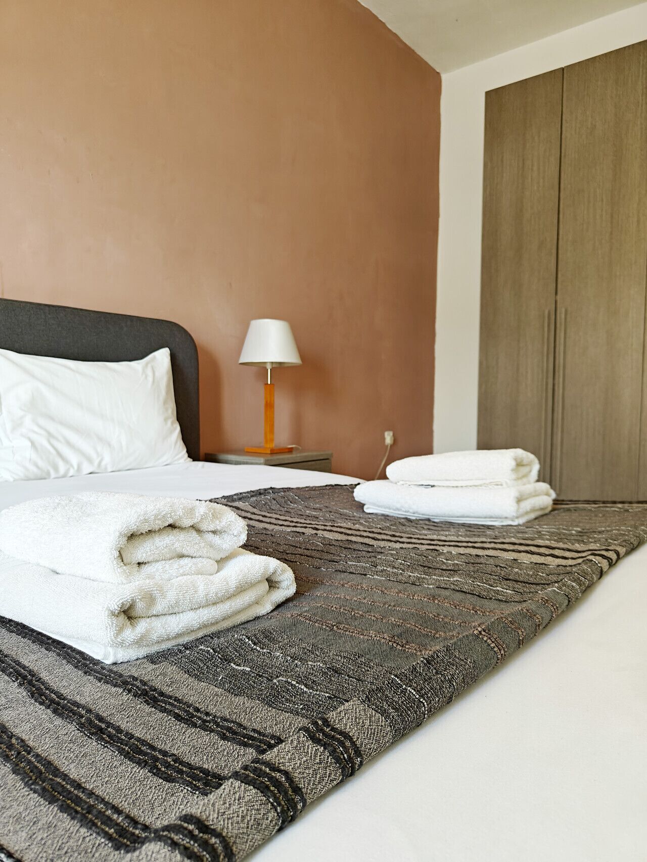 A neatly made bed with white linens and a folded towel, a bedside lamp, and a wardrobe in a hotel room.