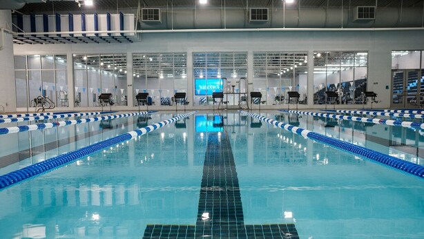 An indoor swimming pool with clear lanes, a digital scoreboard, and a high ceiling with visible structural beams.
