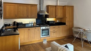 A compact kitchen with wooden cabinets, a gas stove, a microwave, and a dining area with a glass table and chairs. - 1 bedroom accommodation in Falmouth (Falmouth)