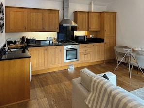 Cozinha compacta com armários de madeira, fogão a gás, micro-ondas e área de refeições com mesa e cadeiras de vidro.