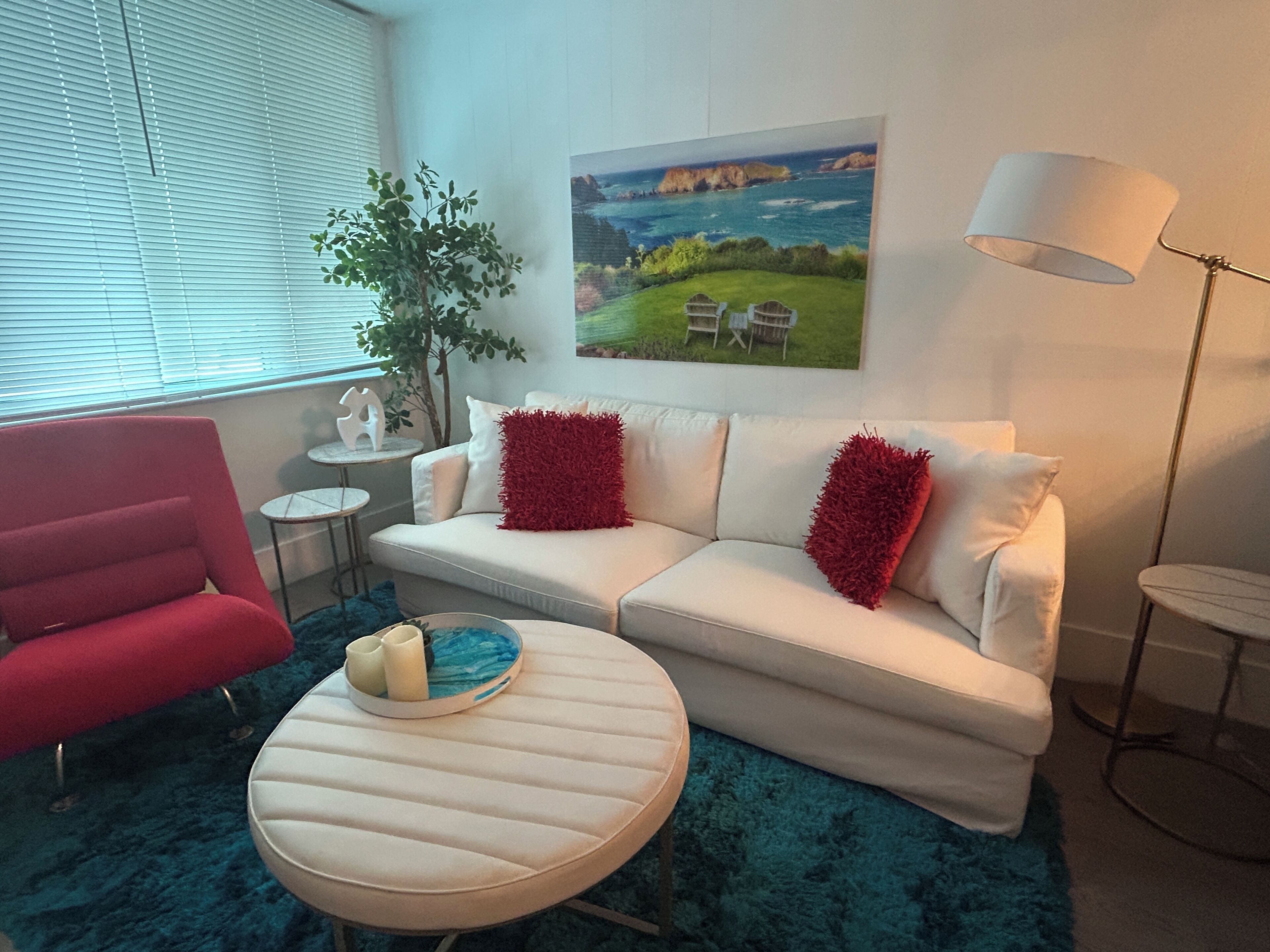 Una sala moderna con un sofá blanco, cojines rojos, una mesa de centro redonda, un sillón rojo, una mesita auxiliar, una lámpara de pie y un cuadro de un paisaje costero.