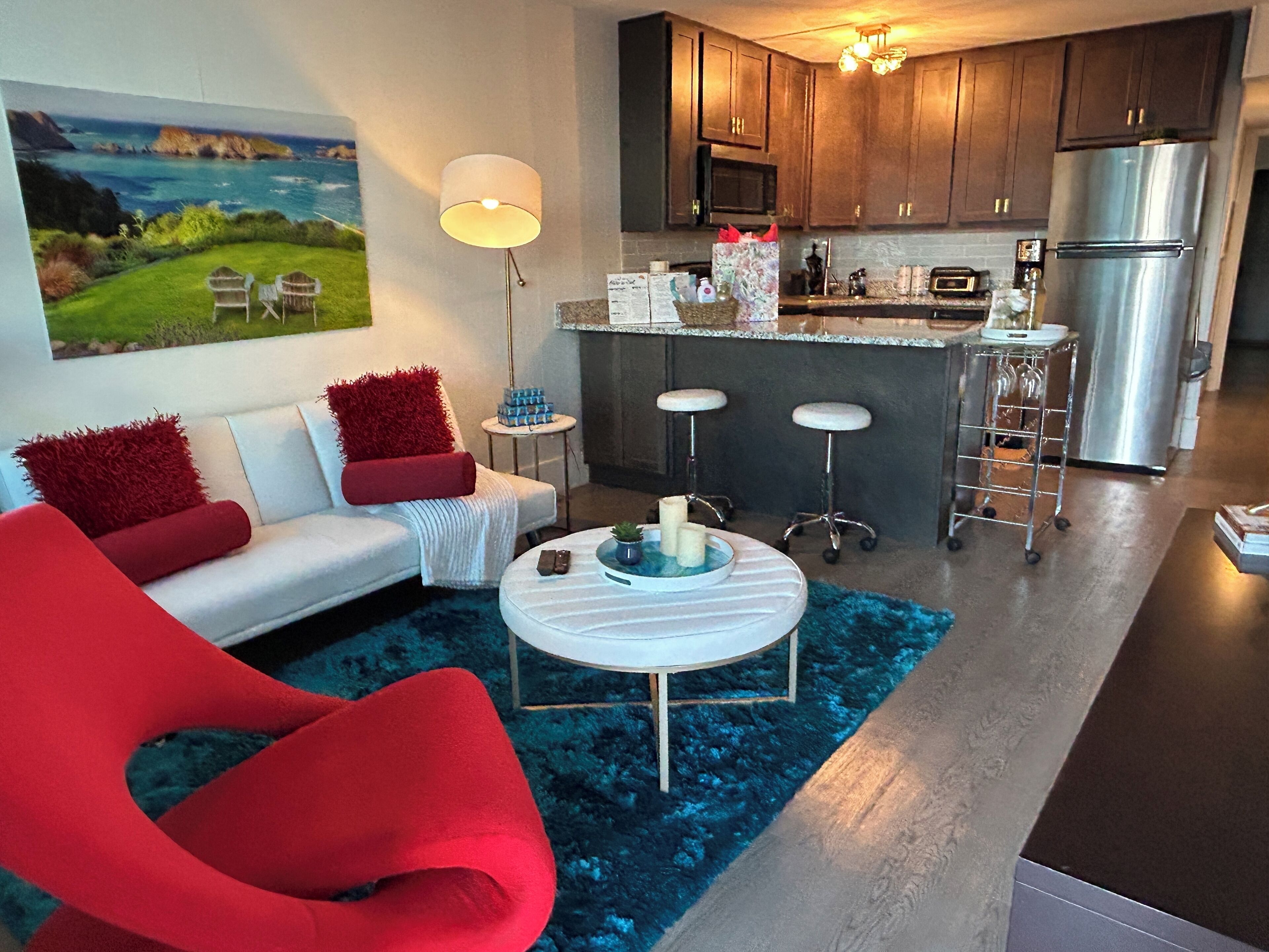 A modern living room with a sofa, red armchair, and a coffee table. There is a kitchen area with a refrigerator, microwave, and bar stools.