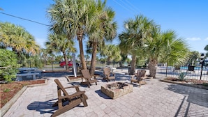 A paved outdoor area with wooden chairs, a fire pit, and palm trees. - Beachside Getaway on Emerald Coast (Fort Walton Beach)