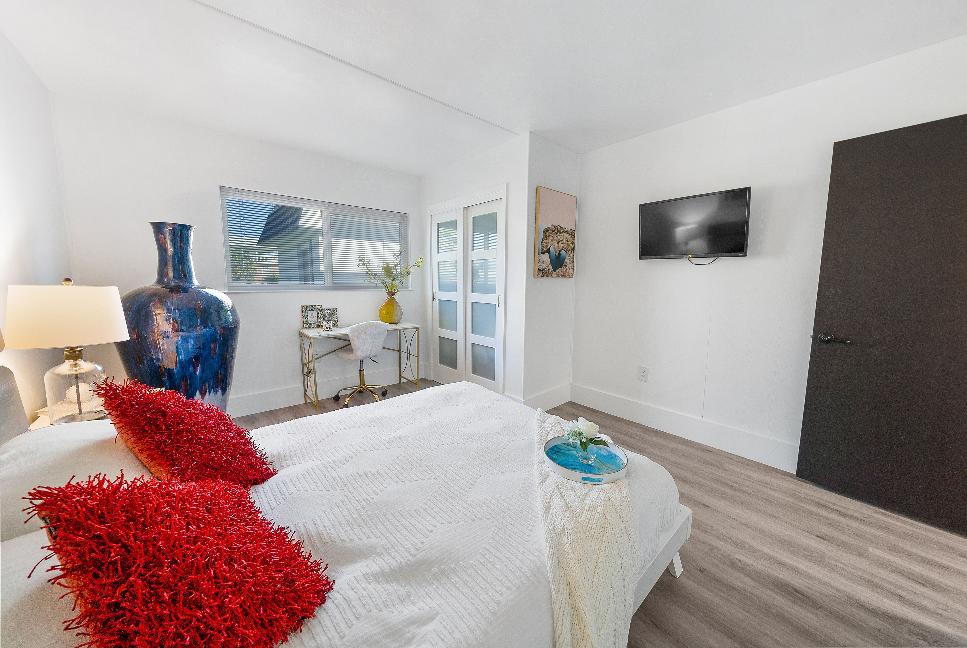 A modern bedroom with a large bed, a TV mounted on the wall, and a window with blinds.