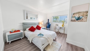 A modern bedroom with a white bed, red pillows, a nightstand, a lamp, a vase, and a framed picture on the wall. - Beachside Getaway on Emerald Coast (Fort Walton Beach)