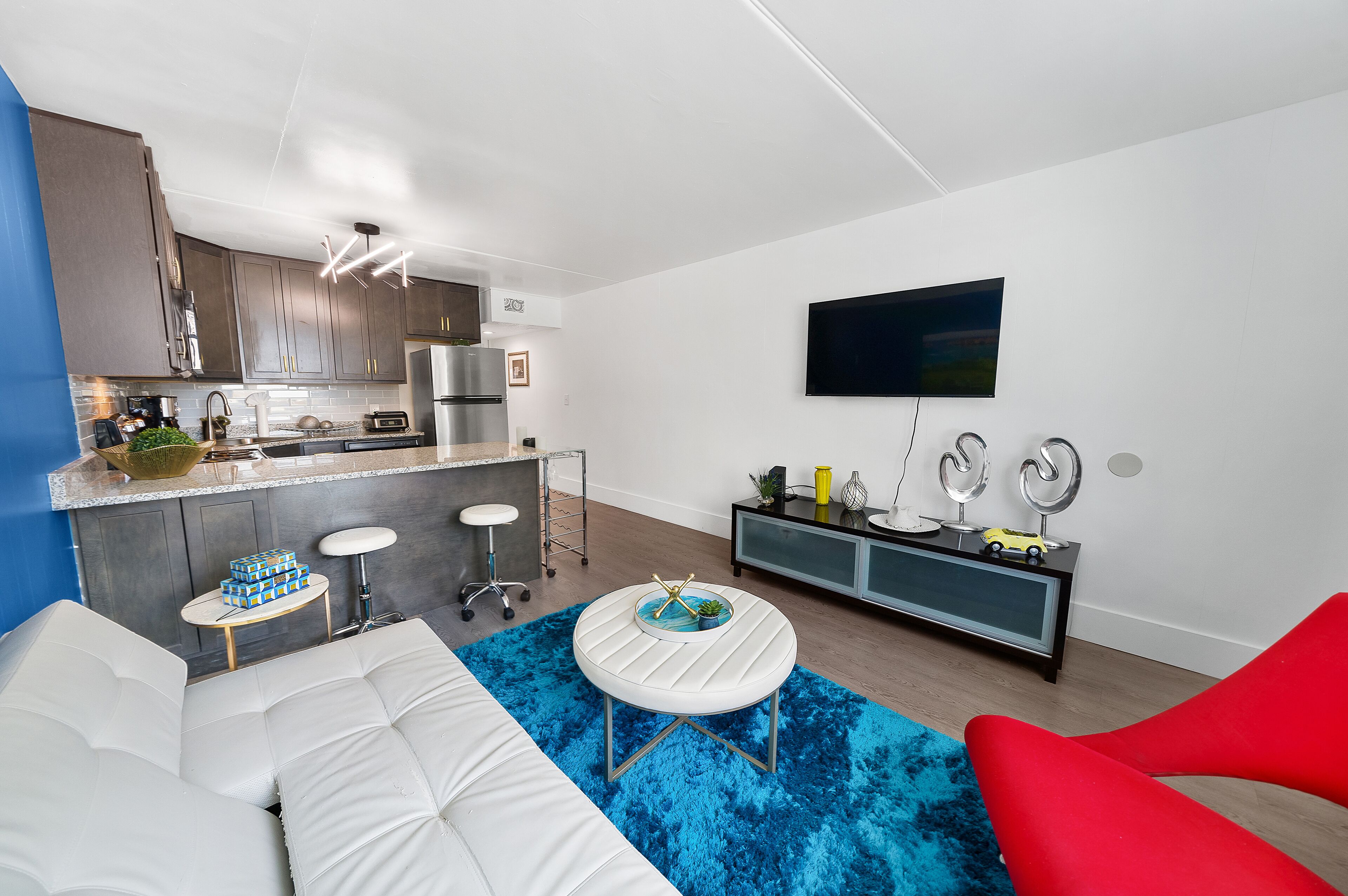 Un salon moderne avec un canapé blanc, un tapis bleu et un fauteuil rouge. Il y a également un coin cuisine équipé d’appareils électroménagers en acier inoxydable et d’un comptoir-bar.
