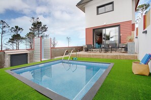 A modern outdoor swimming pool with a glass enclosure, surrounded by artificial grass, and a patio area with outdoor furniture.