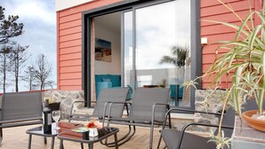 Une terrasse avec des sièges extérieurs, une table avec une bouteille et un verre, et une vue sur l’intérieur du bâtiment.