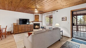 Een gezellige woonkamer met een open haard, houten plafond en een groot raam met zonwering.