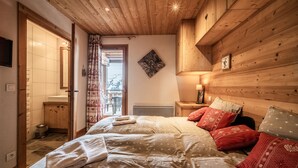 A bedroom with a wooden ceiling, a bed with patterned bedding, a nightstand with a lamp, and a view through an open door. - Apartment "Superb Duplex - Résidence Alpine" with Private Terrace, Balcony and Wi-Fi (Arâches-la-Frasse)