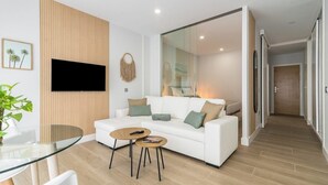 A modern living room with a white sofa, two coffee tables, a TV mounted on the wall, and a glass door leading to a hallway.