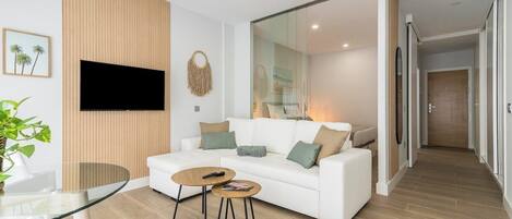 A modern living room with a white sofa, two coffee tables, a TV mounted on the wall, and a glass door leading to a hallway.