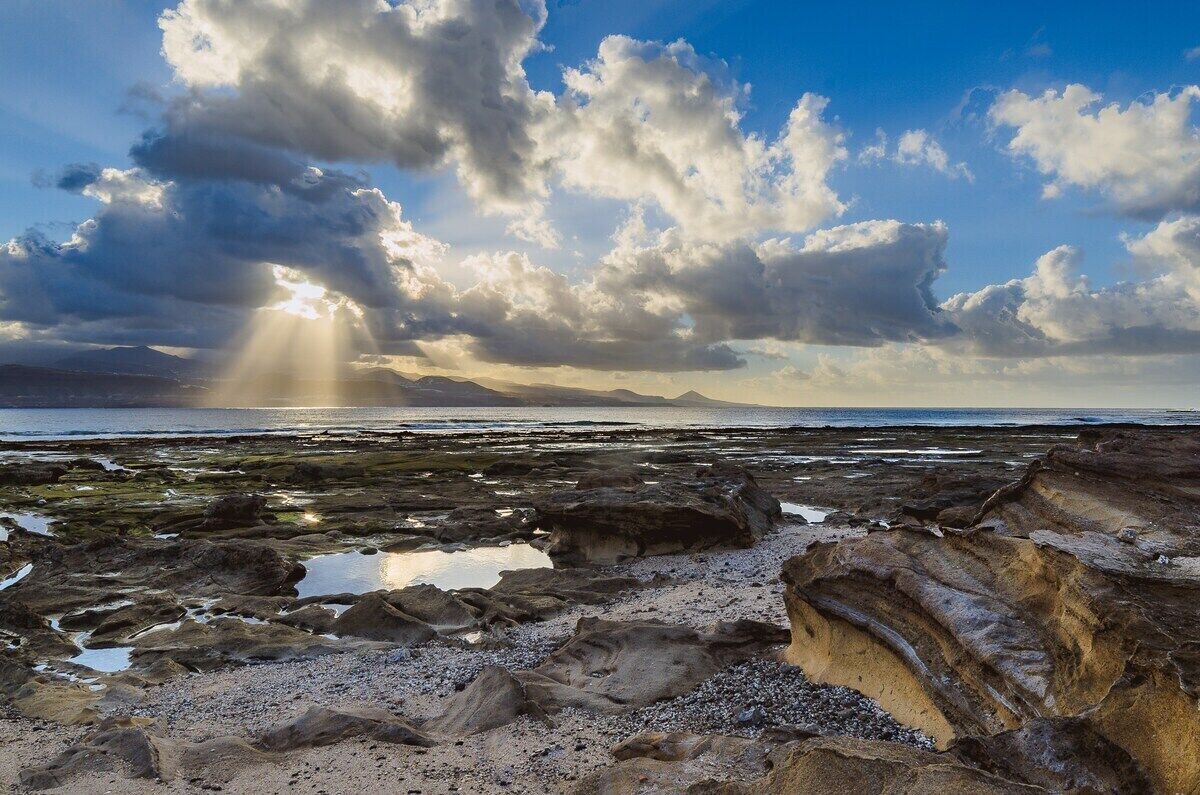 A rocky shoreline with a view of the sea and distant mountains under a partly cloudy sky.