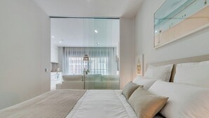 A modern hotel room with a large bed, a glass partition, a dining area, and a view of the city.