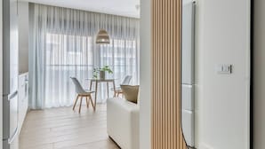 A modern hotel room with a large mirror, wooden paneling, a dining area with a table and chairs, and a kitchen area.