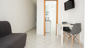 Una sala de estar moderna con un sofá, una mesa de comedor con sillas, un televisor montado en la pared y una cocina visible a través de un vano abierto.