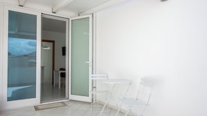 Una habitación con puerta de vidrio corredera que da a un balcón, una pequeña mesa blanca y una silla blanca.