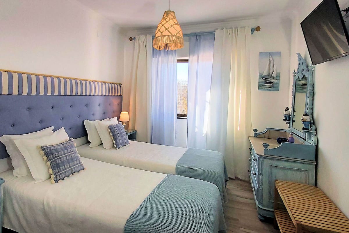 A hotel room with two beds, a wooden bench, a dresser, a television, and a window with curtains.
