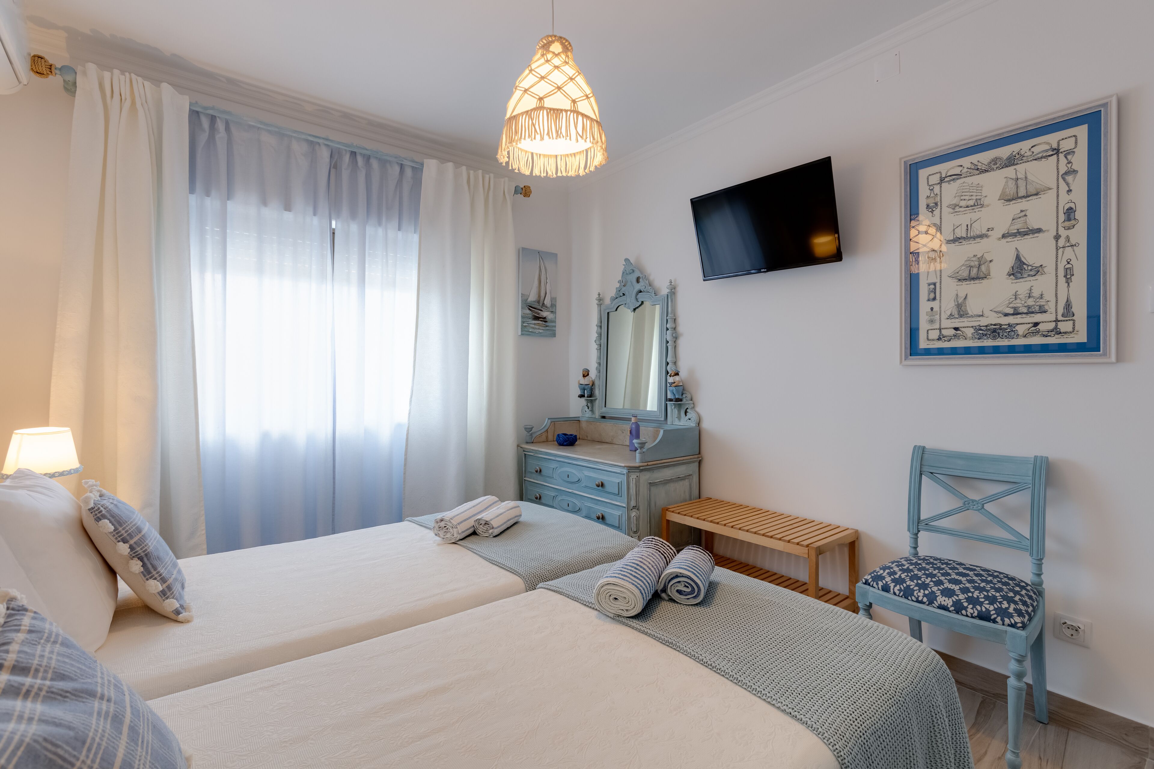 Une chambre d’hôtel avec deux lits, un banc en bois, une chaise, un téléviseur et un tableau encadré au mur.