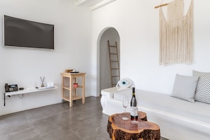 Ein modernes Interieur mit Flachbildfernseher, einer Holzleiter, einer Wanddeko aus Makramee, einer weißen Bank mit gemusterten Kissen und einem Holztisch mit Flasche und Gläsern.