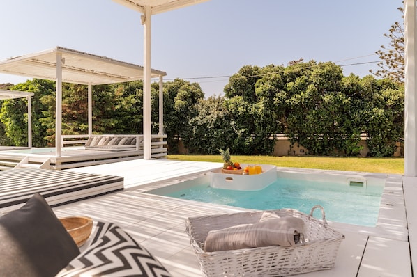 A pool area with lounge chairs, a covered seating area, and a view of a lush garden.