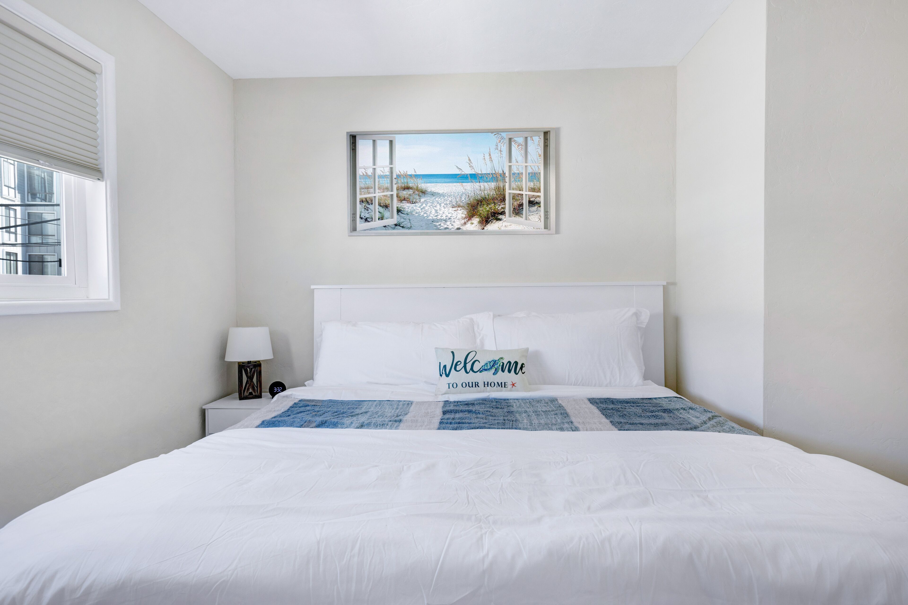 Un dormitorio con cama, mesita de noche, lámpara, ventana con persianas y una pintura con vista al mar.