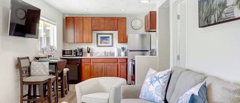 A modern kitchen with wooden cabinets, a stainless steel refrigerator, and a microwave. There is a dining area with a table and chairs, and a living room with a sofa and cushions.
