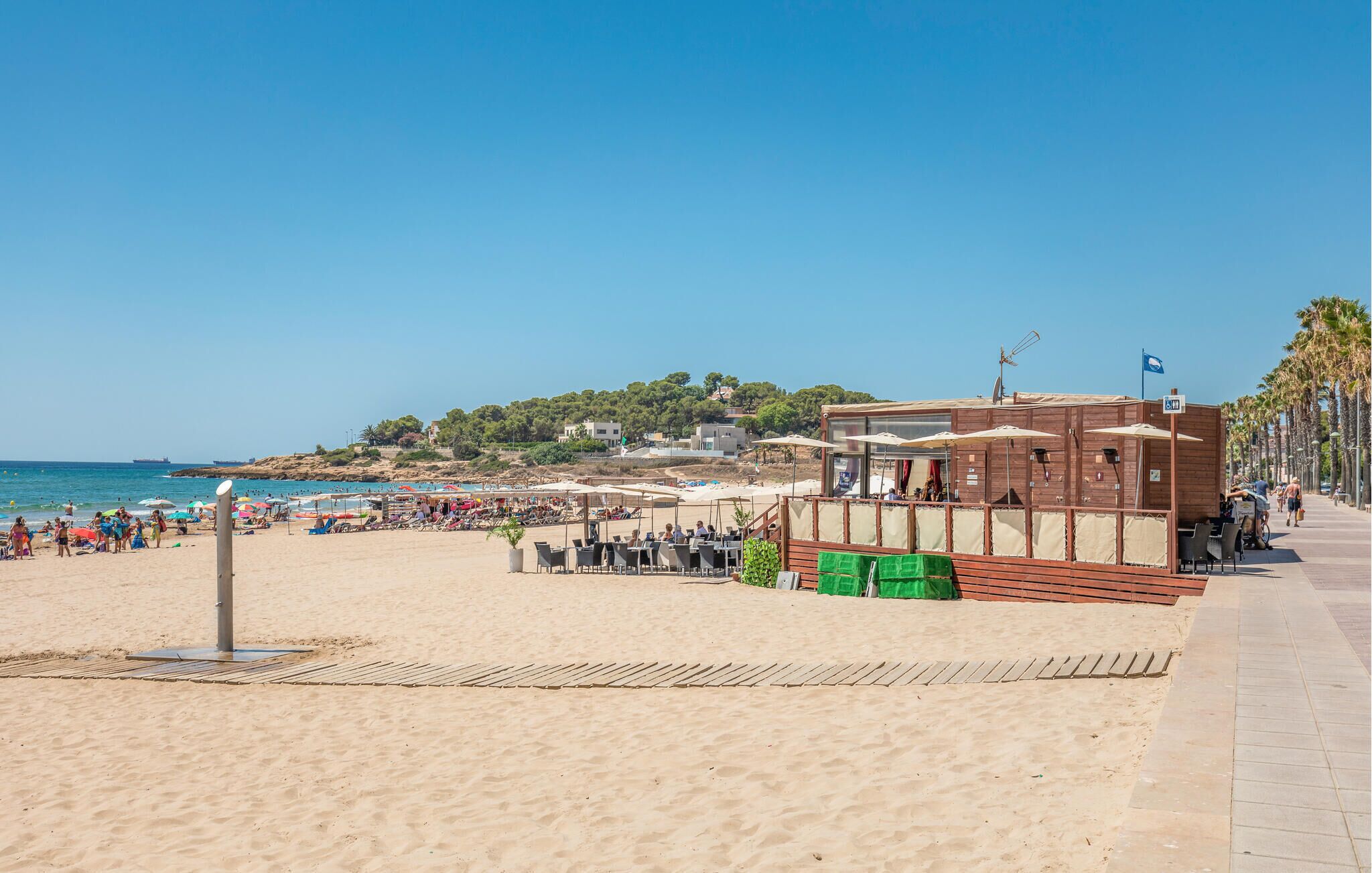 Um restaurante à beira-mar com mesas ao ar livre, uma praia de areia e vista para o mar e colinas ao fundo.