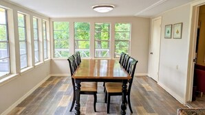 Een eetkamer met een lange houten tafel en stoelen, grote ramen met uitzicht op bomen en een deur met ingelijste foto's.