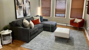 Un salón con un sofá seccional gris, un reposapiés blanco y una alfombra gris. Hay tres ventanas con persianas, un ventilador de techo y un cuadro abstracto grande en la pared.