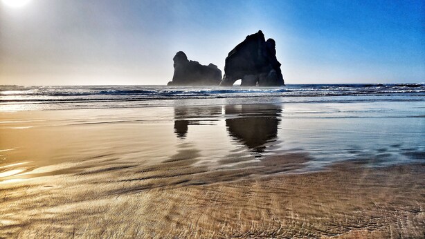 A beach with a large rock formation in the background and calm waves. - Maelstrom End - Eco Farm Stay Cabin (Pūponga)