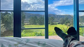 A room with large windows offering a view of a green landscape and mountains. - Maelstrom End - Eco Farm Stay Cabin (Pūponga)