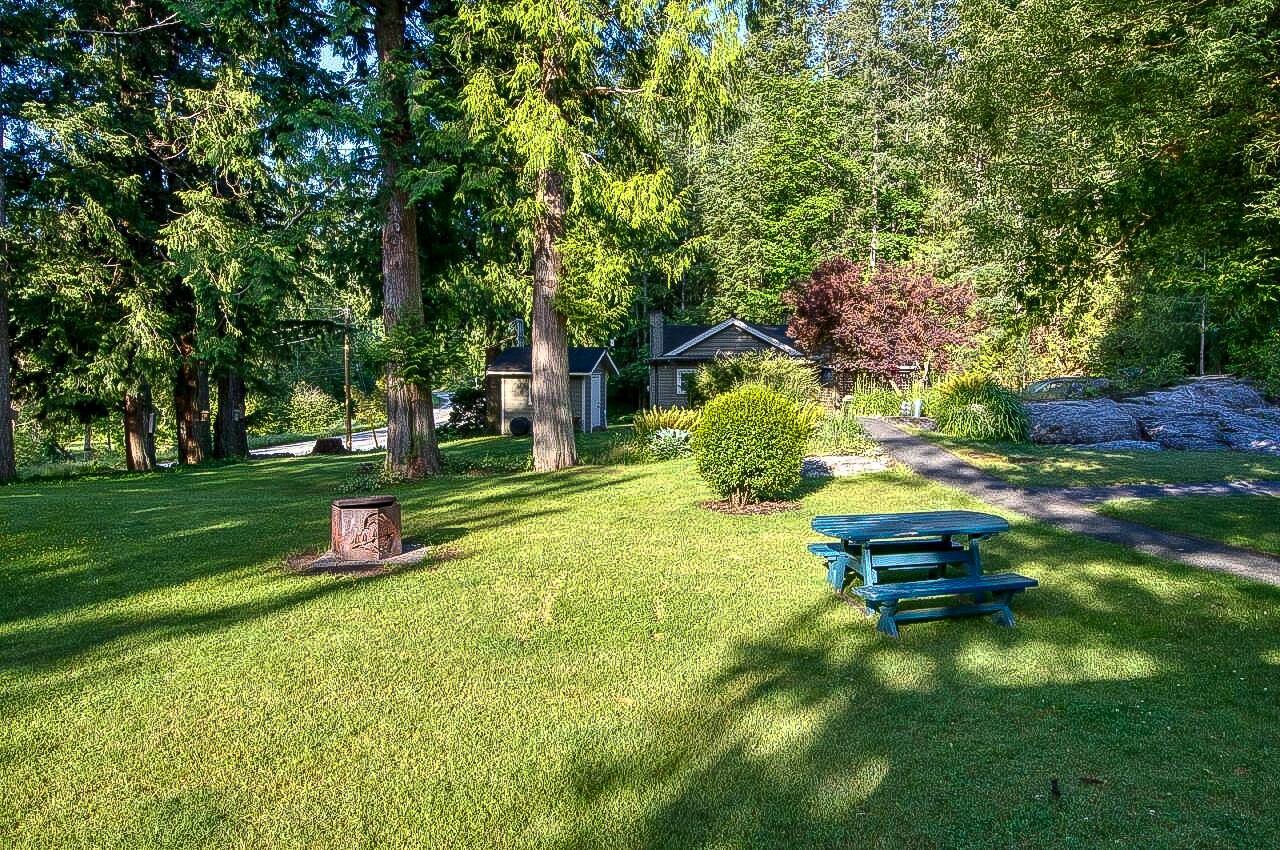 A well-maintained grassy area with a picnic table and a small tree, surrounded by dense trees and a house in the background.