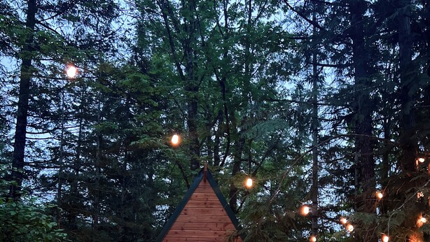 A small wooden cabin with a pitched roof, a bench, a fire pit, and string lights. - The Stonewater (Madeira Park)