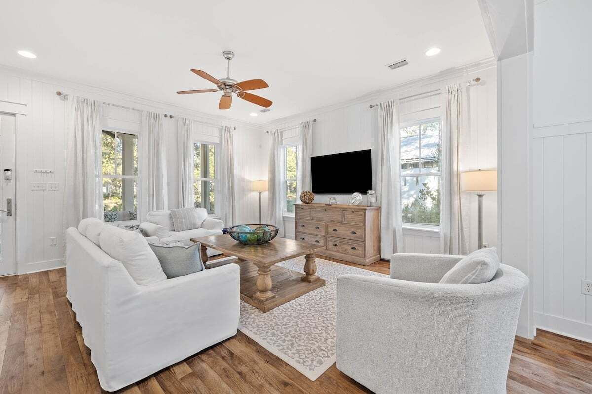 A spacious living room with a ceiling fan, white furniture, a wooden coffee table, and a flat-screen TV.