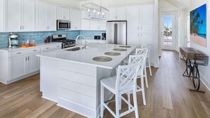 Une cuisine moderne avec des meubles blancs, un grand îlot central doté de tabourets de bar et une vue sur la plage grâce à une porte ouverte.
