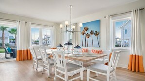 A dining area with a wooden table and white chairs, a chandelier, and a painting of palm trees.
