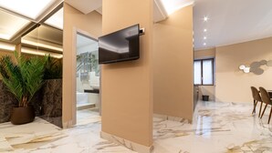 Een moderne hotellobby met een flatscreen televisie aan een muur, marmeren vloer, een potplant en een zithoek met stoelen.