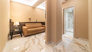Een moderne hotelkamer met een beige sofa, een tafeltje met een lamp, marmeren vloeren en een glazen deur die naar een balkon leidt.