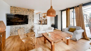 Een moderne woonkamer met een stenen muur, witte banken, een houten salontafel, rieten stoelen en een grote televisie.