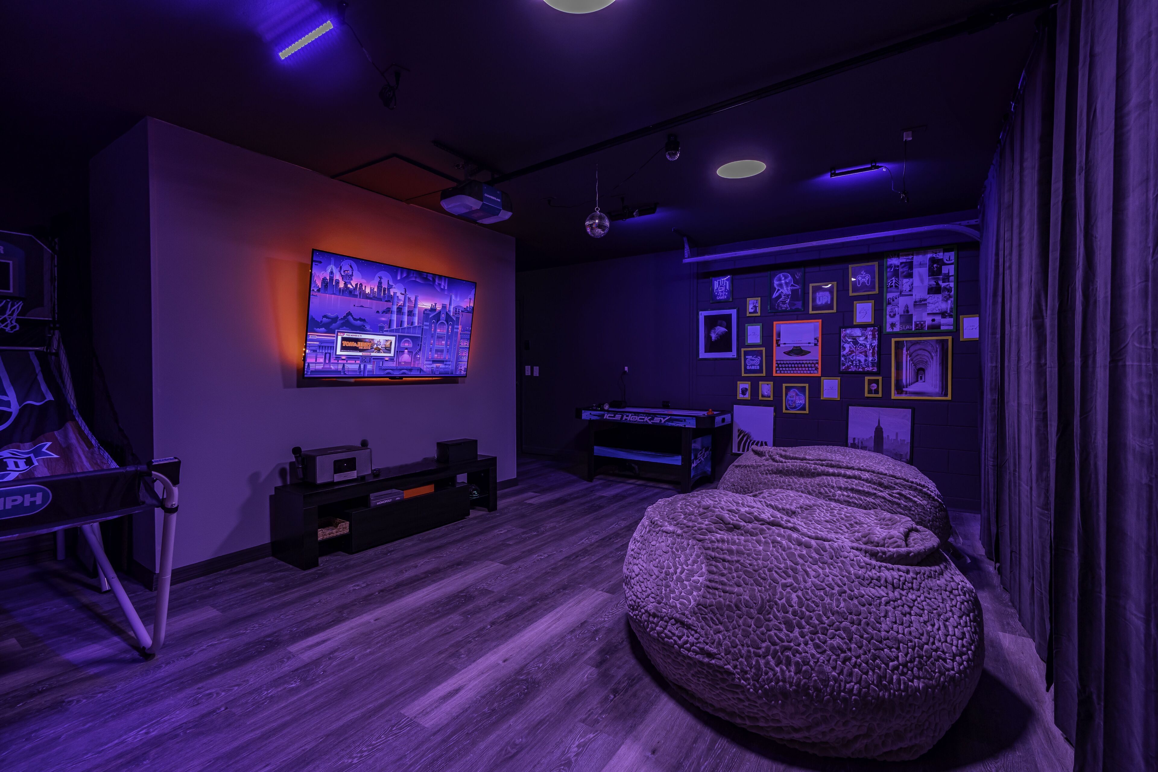 A room with a large flat-screen TV mounted on the wall, a bean bag chair, a pool table, and a wall decorated with framed pictures.