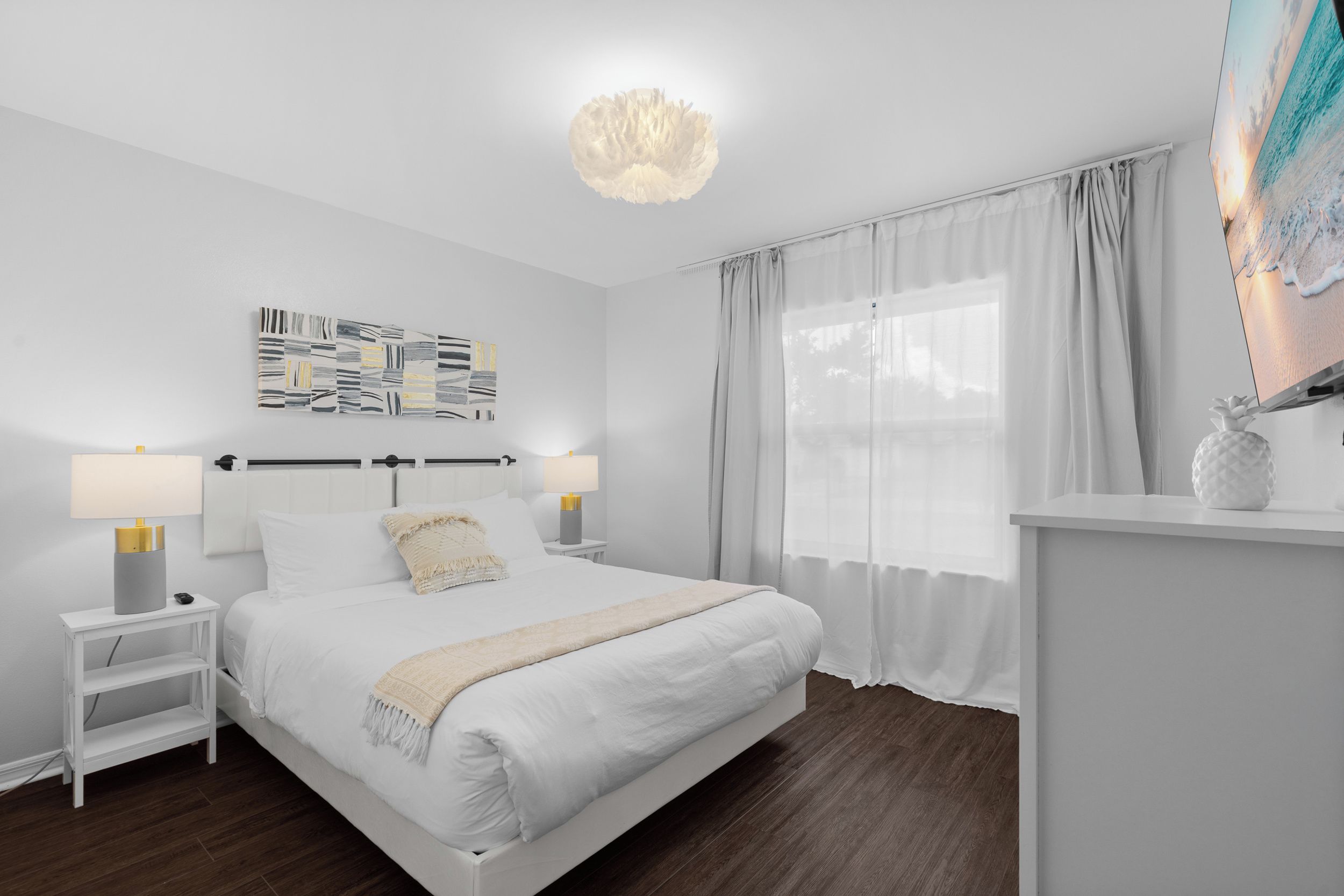 A modern bedroom with a large bed, bedside lamps, a decorative wall piece, and a view of the outdoors through sheer curtains.