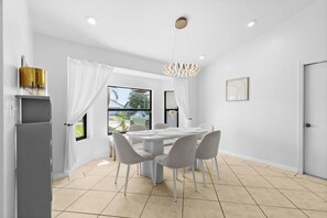 A modern dining room with a white table and chairs, a chandelier, and a view of the outdoors. - Sunshine Villa:3BR Home with Sparkling Heated Pool/Huge Backyard (Davenport)